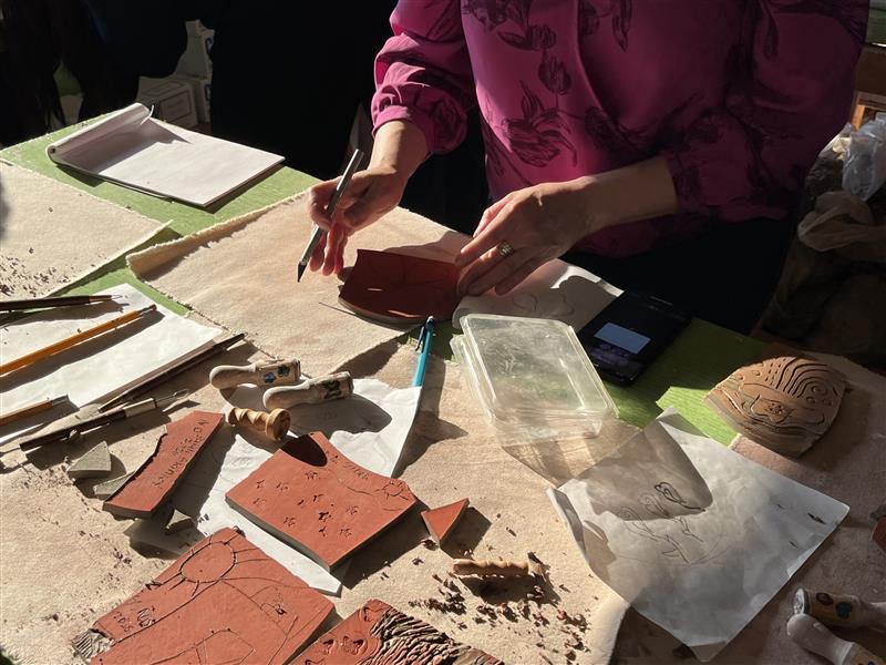 Tile workshop attendee places clay tile onto a canvas mat to begin carving the tile. Around the work station are various tool that will be used to create impressions into the clay tile.