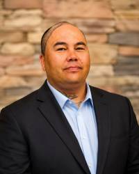 Head and shoulders of Native American Derek Poitra with a black suit jacket, collared under-shirt, closed half-smile, very short hair that may be pulled back, with a blurred brick background
