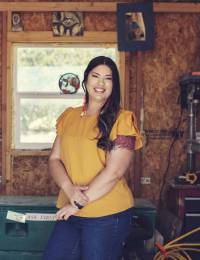 Knees to head image of (brightly smiling) Native American Shayna Grandbois-Herrera with long black hair, wearing Native jewelry, bright colors, and jeans, with her Native artwork behind her
